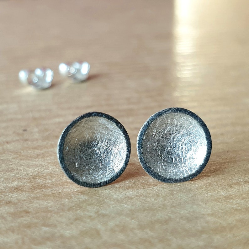 May include: A pair of silver earrings with a textured, circular design. The earrings have a dark gray rim.