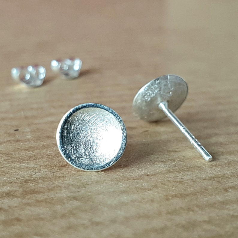 May include: Two silver earring studs, one with a round, flat, textured surface and the other with a post and a backing.
