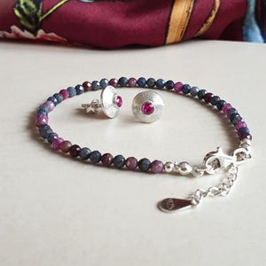 Könnte beinhalten: Ein Schmuckset mit einem Perlenarmband und passenden Ohrsteckern. Das Armband und die Ohrringe bestehen aus kleinen, facettierten Perlen in Blau-, Lila- und Rottönen. Die Ohrringe und der Armbandverschluss sind silberfarben, mit einem leuchtend rosa Mittelstein.