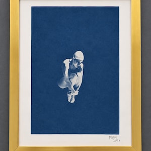 May include: A framed cyanotype print featuring a white silhouette of a swimmer in a tuck position against a deep blue background. The artwork is enclosed in a metallic gold frame and includes the handwritten signature B2S 2024 at the bottom.