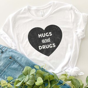 May include: White t-shirt with a black heart graphic that says "Hugs and Drugs" in a vintage font.