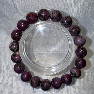Peut inclure: Un bracelet en pierres précieuses de couleur pourpre et noire avec des perles rondes. Le bracelet est sur un présentoir transparent.