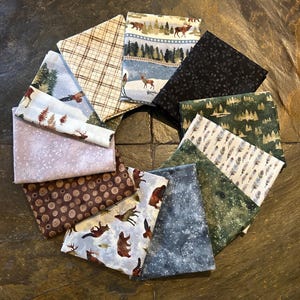 May include: A collection of 10 fabric squares featuring a woodland theme. The fabrics are a mix of patterns and colors, including brown, green, blue, and white. Some of the patterns include trees, deer, bears, and snowflakes.