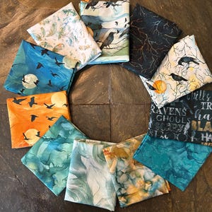 May include: A collection of ten folded fabric squares arranged in a circle. The patterns include ravens, moons, bare tree branches, abstract teal and gold ink swirls, and text reading RAVENS, GHOULS, SCARY, and BOO in various fonts and colors.