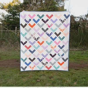 May include: A white quilt with a repeating chevron pattern in various colors, including pink, blue, orange, and gray. The quilt is labeled "The Freya Quilt" and is designed by Erica Jackman. The quilt is designed for baby, lap, and twin sizes.
