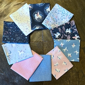 May include: A collection of fabric squares with various patterns, including unicorns, stars, and floral designs. The color palette includes shades of blue, pink, and white. The fabrics are arranged in a circular pattern on a dark surface.