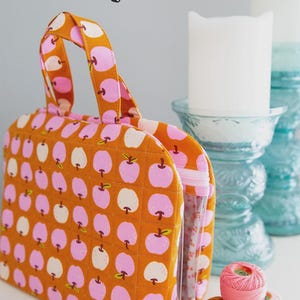 May include: A quilted orange tote bag with a repeating pattern of pink and white apples. The bag has a clear pocket and two handles. The text "THE TRUVY BAG" is at the top. The logo "KNOT + THREAD design" is at the bottom.