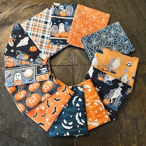 May include: A collection of Halloween-themed fabric squares arranged in a circular pattern. The fabrics feature various designs, including pumpkins, ghosts, bats, spiderwebs, and tombstones. Colors include orange, black, white, and blue.