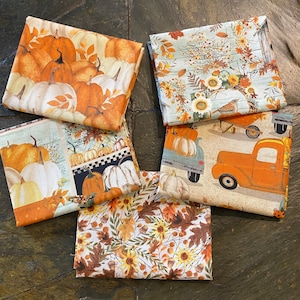 May include: Five folded fabric squares with autumn-themed patterns. Designs include pumpkins, sunflowers, leaves, and vintage trucks in shades of orange, white, and brown. The fabrics are arranged on a dark, textured surface.