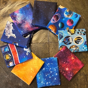 May include: A collection of square fabric pieces with space-themed designs. The fabrics feature images of galaxies, planets, astronauts, and constellations in shades of blue, purple, orange, and white. The pieces are arranged in a circular pattern.