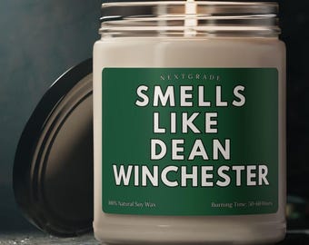 Op de afbeelding: Een brandende kaars in een glazen pot met een zwart deksel. Het etiket leest "SMELLS LIKE DEAN WINCHESTER" in witte tekst op een groene achtergrond. De kaars is gemaakt met 100% natuurlijke sojawas en heeft een brandtijd van 50-60 uur.