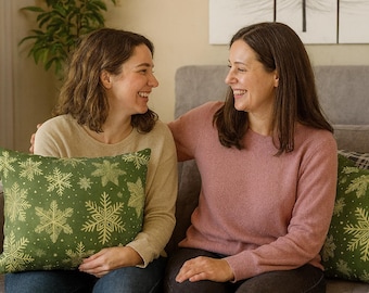 May include: Two women smiling, sitting on a gray couch. One woman holds a green pillow with gold snowflake designs. The other woman wears a pink sweater. A green plant is in the background.