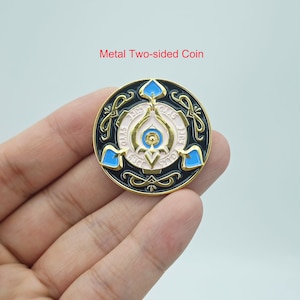 May include: A gold metal coin with a black background and blue and pink accents. The coin features a stylized design with the words "THIS TRY" and "GALS LIKE" on the top and bottom of the design. The coin is being held in a hand.