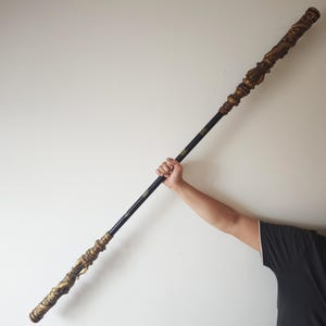 May include: A long, black and gold staff with intricate details. The staff is held by a person's hand, with the rest of the person's arm and torso visible.