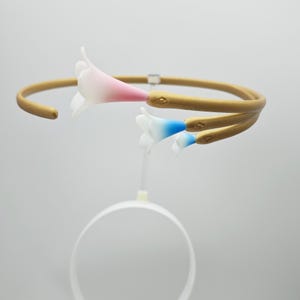 Puede incluir: Un brazalete dorado con tres delicados abalorios de flores blancas. Las flores tienen un degradado de rosa, azul y blanco.