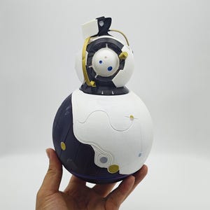 May include: A white and black spherical robot with a gold accent and a blue eye. The robot has a smaller, round head with a black and white design. The robot is being held in a hand.