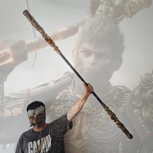 May include: A person holding a long, black and gold staff with intricate details. The staff is decorated with gold accents and has a pointed tip. The staff is held in front of a mural depicting a monkey warrior with a golden crown and armor.