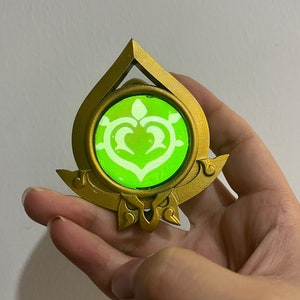 May include: A gold-coloured pendant featuring a green, glowing, heart-shaped symbol within. The pendant is shaped like a teardrop and has intricate details.