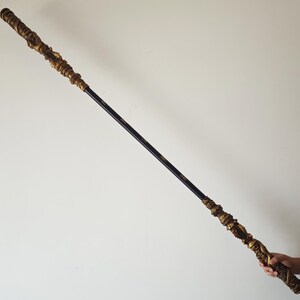 May include: A long, black and gold staff with intricate details. The staff is held by a hand in the lower right corner of the image.