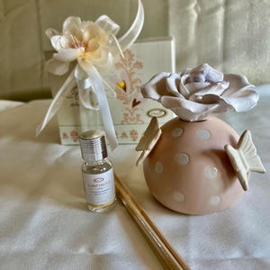 Puede incluir: Juego de fragancia para el hogar decorativo. Incluye un difusor de cerámica rosa claro con lunares blancos y detalles de mariposas, una pequeña botella de vidrio con "L'Infinito 1972", un paquete de cañas y una caja de regalo floral.