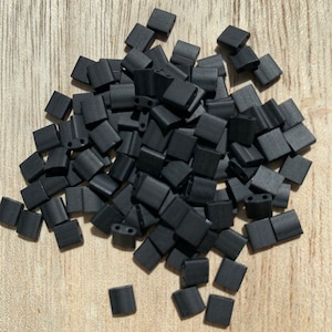 Könnte beinhalten: Ein Haufen schwarzer quadratischer Perlen. Die Perlen sind aus mattem Material und haben jeweils zwei Löcher.