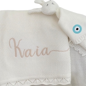 May include: White knitted baby blanket with a crocheted border and a pink embroidered name "Kaia". A white bunny toy with a blue evil eye charm is attached to the blanket.