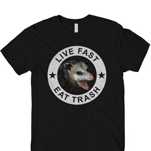 May include: Black t-shirt with a white circle graphic featuring a possum with its mouth open and the text "Live Fast Eat Trash".