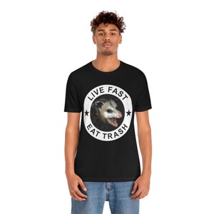 May include: Black t-shirt with a white circle graphic featuring a possum with the text "Live Fast Eat Trash".