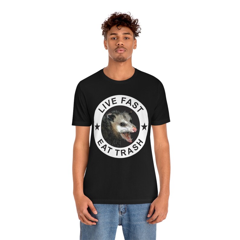 May include: Black t-shirt with a white circle graphic featuring a possum with the text "Live Fast Eat Trash".