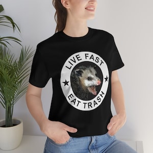 May include: Black t-shirt with a white circle graphic featuring a possum and the text "Live Fast Eat Trash".