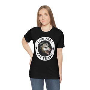 May include: A black t-shirt with a white circle graphic featuring a possum with the text "Live Fast Eat Trash".