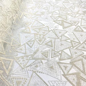 May include: Ivory-colored fabric with a repeating geometric triangle pattern. The triangles are outlined with a textured, possibly beaded, material, and some are filled with intricate designs. The fabric appears to be delicate and suitable for special occasion wear.