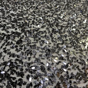 May include: Close-up of a black fabric with a net-like texture, densely covered in small, shiny, leaf-shaped sequins. The sequins catch the light, creating a sparkling effect. The overall appearance is dark and elegant.