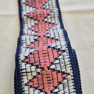 Könnte beinhalten: Ein gewebtes Zierband mit einem sich wiederholenden geometrischen Muster in Rosa, Weiß und Marineblau. Die Zierleiste ist etwa 5 cm breit und hat eine strukturierte Oberfläche. Das Design ist eine Reihe von Rauten.