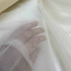 Könnte beinhalten: Nahaufnahme von durchsichtigem, elfenbeinfarbenem Tüllstoff. Der Stoff ist sehr fein und zart, mit einer weichen, fließenden Textur.