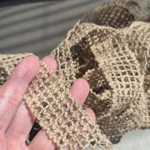 May include: Close-up of a section of tan-colored, open-weave jute ribbon. The ribbon has a grid-like pattern and is held in a hand. A ruler and a tag with text are partially visible.