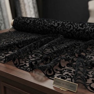 May include: A roll of black sheer fabric featuring a raised velvet-like scrollwork pattern. The material is draped across a surface, and a small rectangular sign with the text FABRIC BY THE YARD is placed in the foreground of the display.