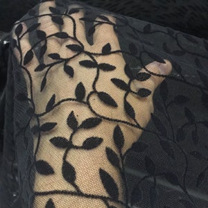 Könnte beinhalten: Ein transparenter schwarzer Stoff mit einem zarten, sich wiederholenden Ranken- und Blattmuster in samtiger Textur. Das Material ist halbtransparent und weist eine feine Netzstruktur auf, die Licht durch die Lücken zwischen dem dunklen Blattwerk lässt.