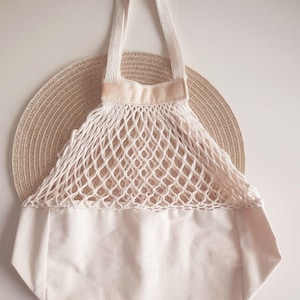 Puede incluir: Bolsa de compras de red blanquecina con base de lona y asas a juego. La bolsa está colocada sobre un fondo beige tejido. El diseño de la red es abierto y la bolsa parece estar vacía.