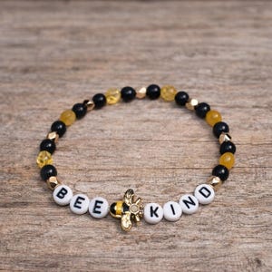 Pode incluir: Uma pulseira de contas pretas, douradas e amarelas. A pulseira apresenta contas de letras brancas que soletram "BEE KIND" com um berloque de abelha dourada no centro. A pulseira está em uma superfície de madeira.