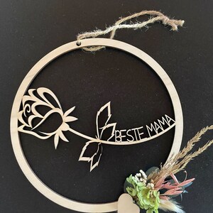 Könnte beinhalten: Ein Holz-Kreis mit einer ausgeschnittenen Rose und dem Text "Beste Mama" darauf. Ein kleines Herz und getrocknete Blumen sind am unteren Rand des Kreises befestigt.