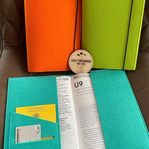 Könnte beinhalten: Ein blauer Filzordner mit einer gelben Karte und einer weißen Karte im Inneren. Der Ordner hat ein schwarzes Gummiband und eine Tasche auf der Vorderseite. Der Ordner ist geöffnet und zeigt ein Buch mit dem Text "U9 Elterninformation Untersuchung vor 60. bis zum 64, 1." Das Buch liegt auf einer braunen Oberfläche. Im Hintergrund befindet sich ein Holzschild mit der Aufschrift "WOID CUSTOMMADE SINCE 2022".