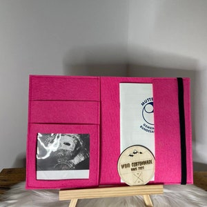 Könnte beinhalten: Ein rosa Filzordner mit einem schwarzen Gummibandverschluss. Der Ordner hat ein transparentes Plastikfenster, in dem ein Schwarzweiß-Ultraschallbild zu sehen ist. Ein Holz-Kreis mit dem Text "VOID CUSTOMMADE SINCE 2022" ist am Ordner befestigt.