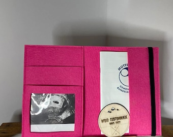 Könnte beinhalten: Ein rosa Filzordner mit einem schwarzen Gummibandverschluss. Der Ordner hat ein transparentes Plastikfenster, in dem ein Schwarzweiß-Ultraschallbild zu sehen ist. Ein Holz-Kreis mit dem Text "VOID CUSTOMMADE SINCE 2022" ist am Ordner befestigt.