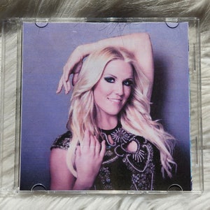 May include: A CD case showing a woman with long blonde hair, wearing a black top with decorative embellishments. The image has a purple background and is set against a white, fluffy surface.