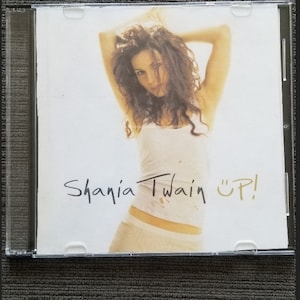 May include: A CD case with a white background featuring a woman with long brown hair wearing a white vest top. The text "Shania Twain "UP!" is written in black on the bottom right corner of the case.