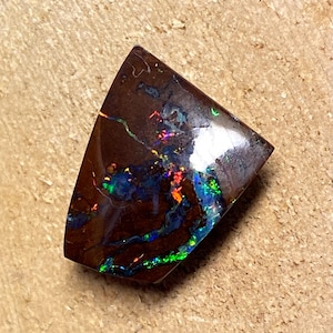 May include: A dark brown, iridescent opal with flashes of green, blue, and red. The opal is cut into a teardrop shape and has a smooth, polished surface.