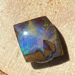 Könnte beinhalten: Ein dunkelbrauner, schwarzer und blauer Opal-Edelstein mit einer rauen, unregelmäßigen Form. Der Opal hat einen hellen, irisierenden Glanz.