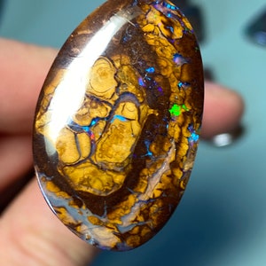 May include: An oval-shaped opal gemstone in brown with a white, iridescent sheen and flashes of blue, green, and purple.