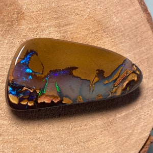 May include: A brown, blue, and green opal cabochon with a rough, natural shape. The opal has a dark brown base with flashes of blue, green, and purple.